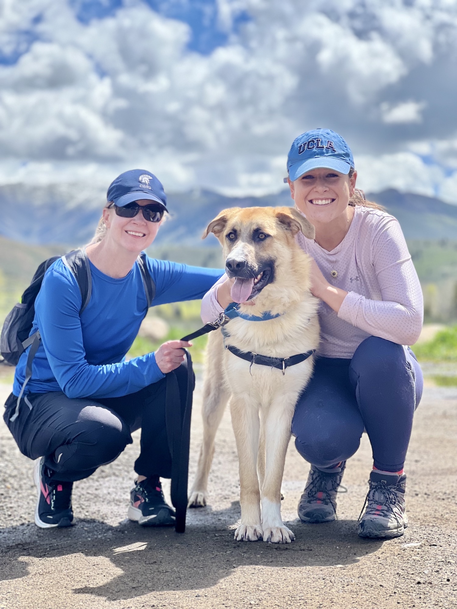 Two women in hiking gear squatting down on either side of a large dog all smiling at the camera