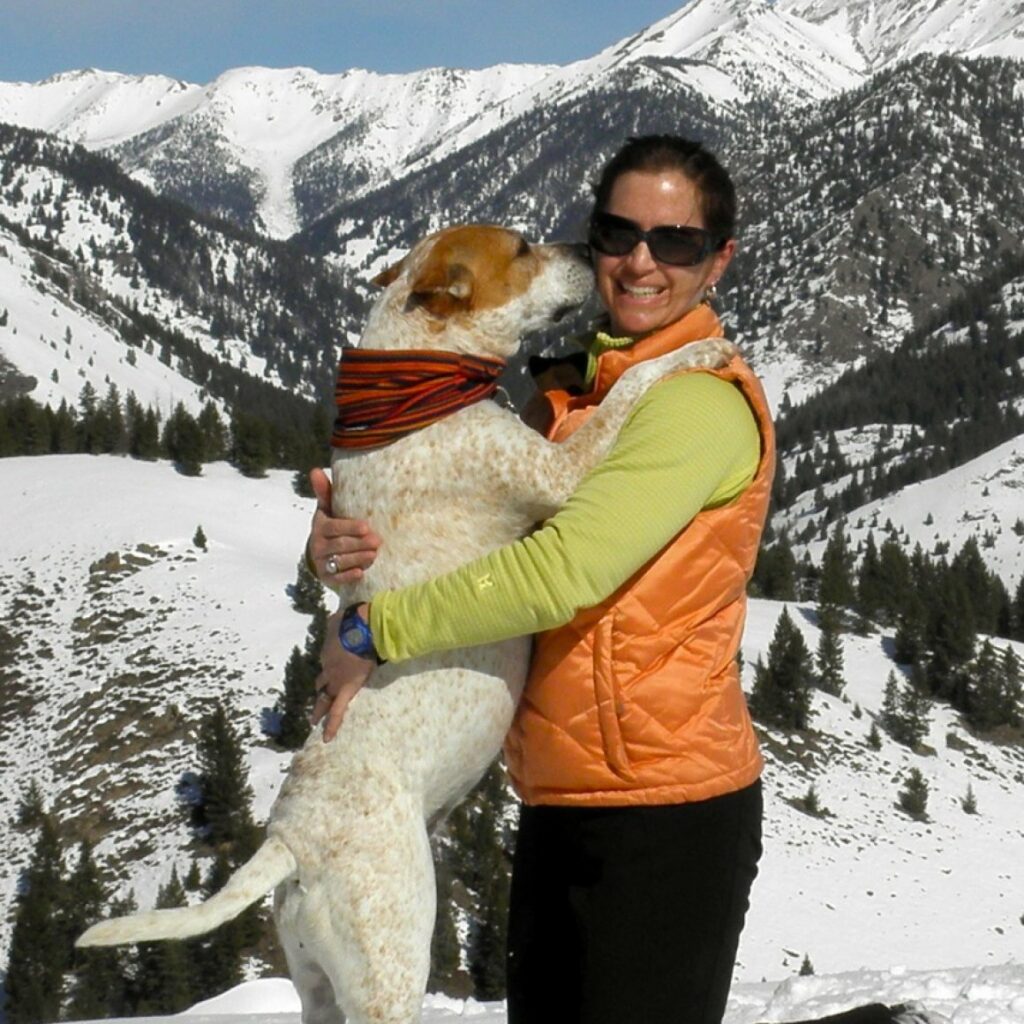 Stephanie Eisenbarth hugging her dog who is standing on two legs, hugging her back, with a snowy mountain background