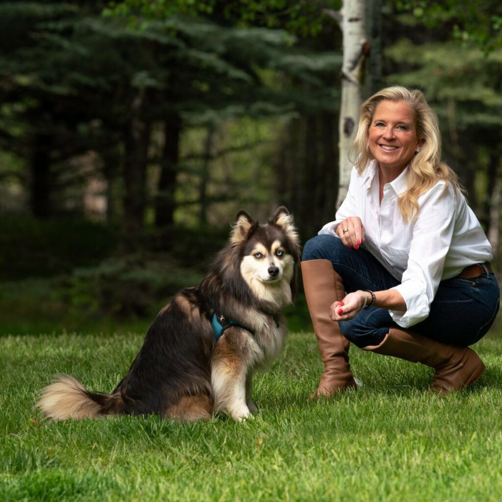 Corry Hart Clayville crouching next to her dog on bright green grass in front of a forested area