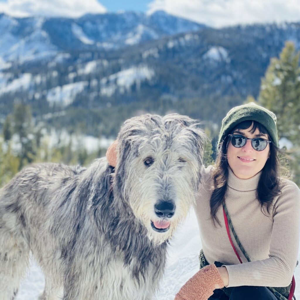 Kelsey Strahle kneeling down with an arm around her large Irish Wolfhound in front of a snowy mountain landscape