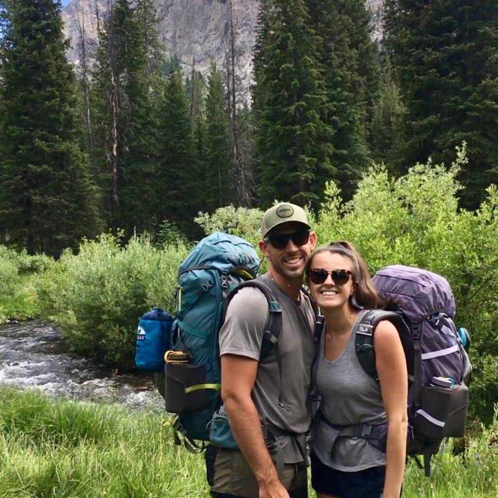Lauren and her husband, Justin, in hiking gear with large backpacks posing for a photo together in front of a river and evergreen trees